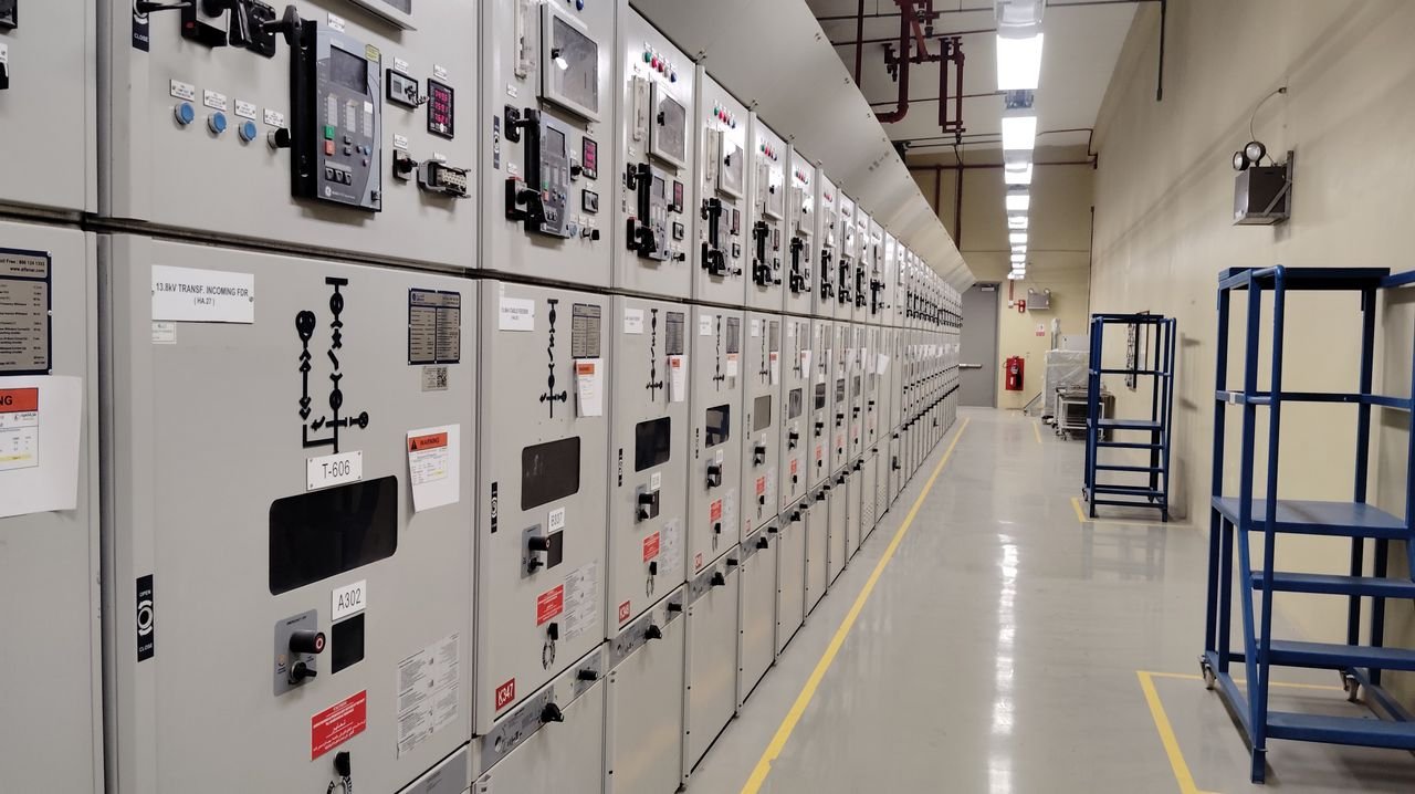 Long row of control panels in an industrial electrical control room, well-lit and spacious.